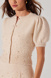 A woman is wearing a cream-colored, short-sleeved knit cardigan with a round neckline. The cardigan features small pearl embellishments scattered across the front and around the waist, with a row of larger pearls at the hem. The sleeves are puffed with ribbed cuffs.